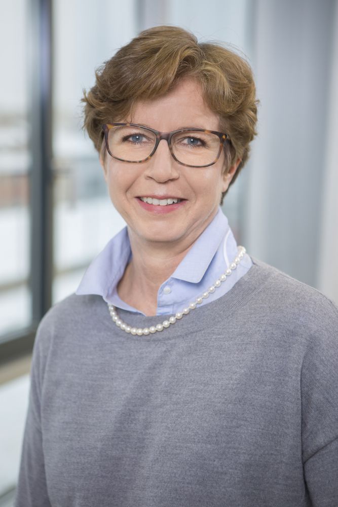 Prof. Dr. med. Elke Holinski-Feder, seriös lächelnd, trägt einen grauen Pullover über einer blauen Bluse und Brille